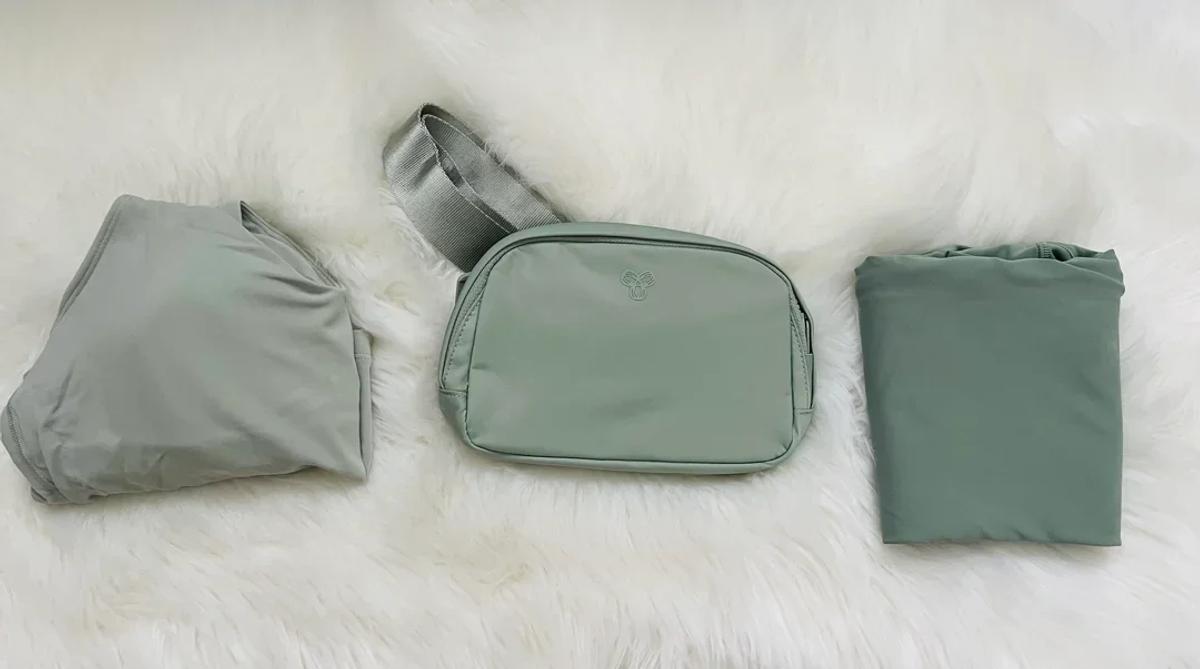 Three folded grey green nylon items laid on a white fur surface: a packed bag, a small zipped pouch and a folded garment.