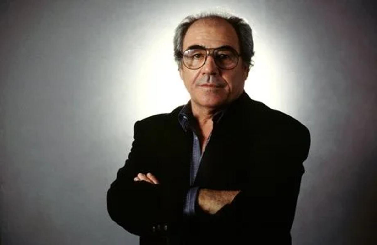 A portrait of a man with grey hair and glasses in a dark jacket, arms folded, lit against a dark background.