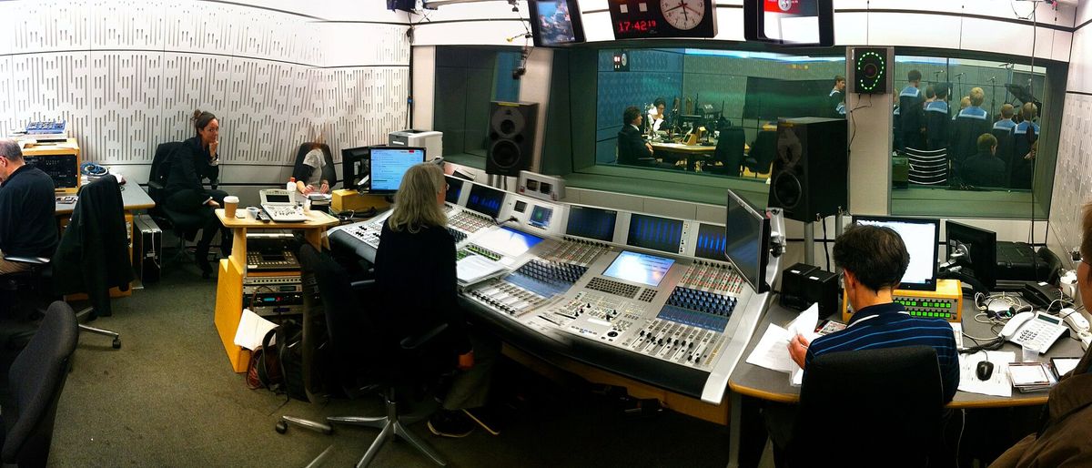 A broadcast control room with a large mixing desk, monitors, a studio clock and a glass window onto the studio beyond.