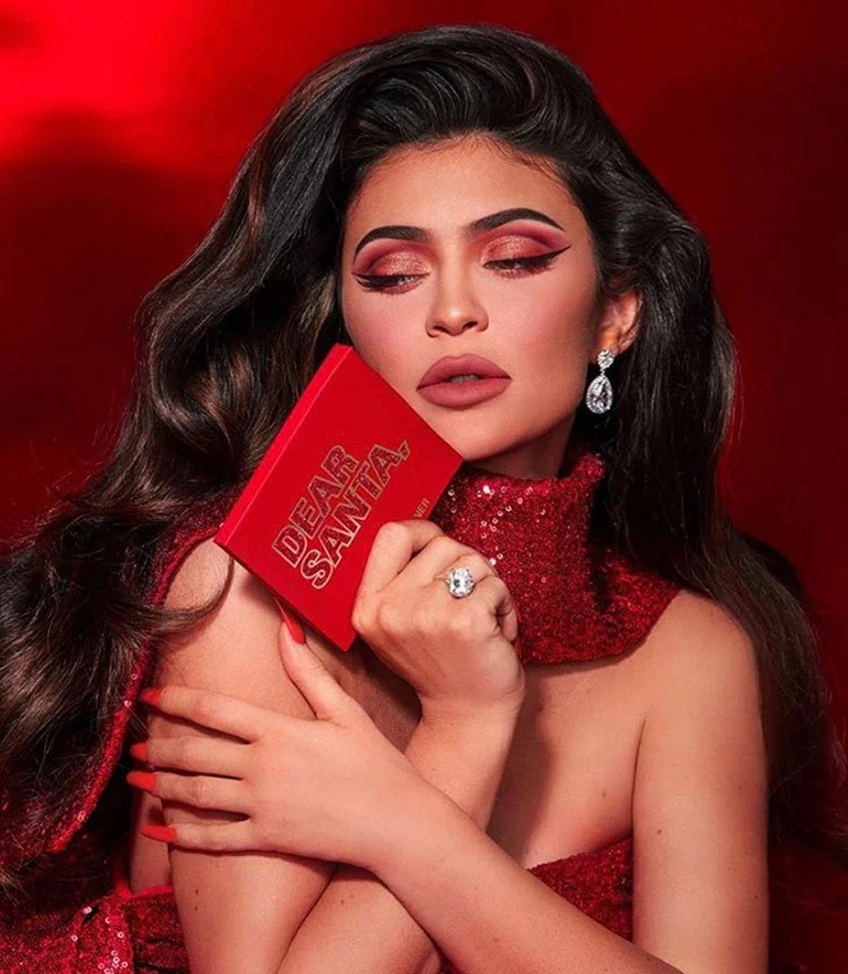 A woman in a red sequinned dress against a red background, hair styled in waves, holding a red book to her chest.