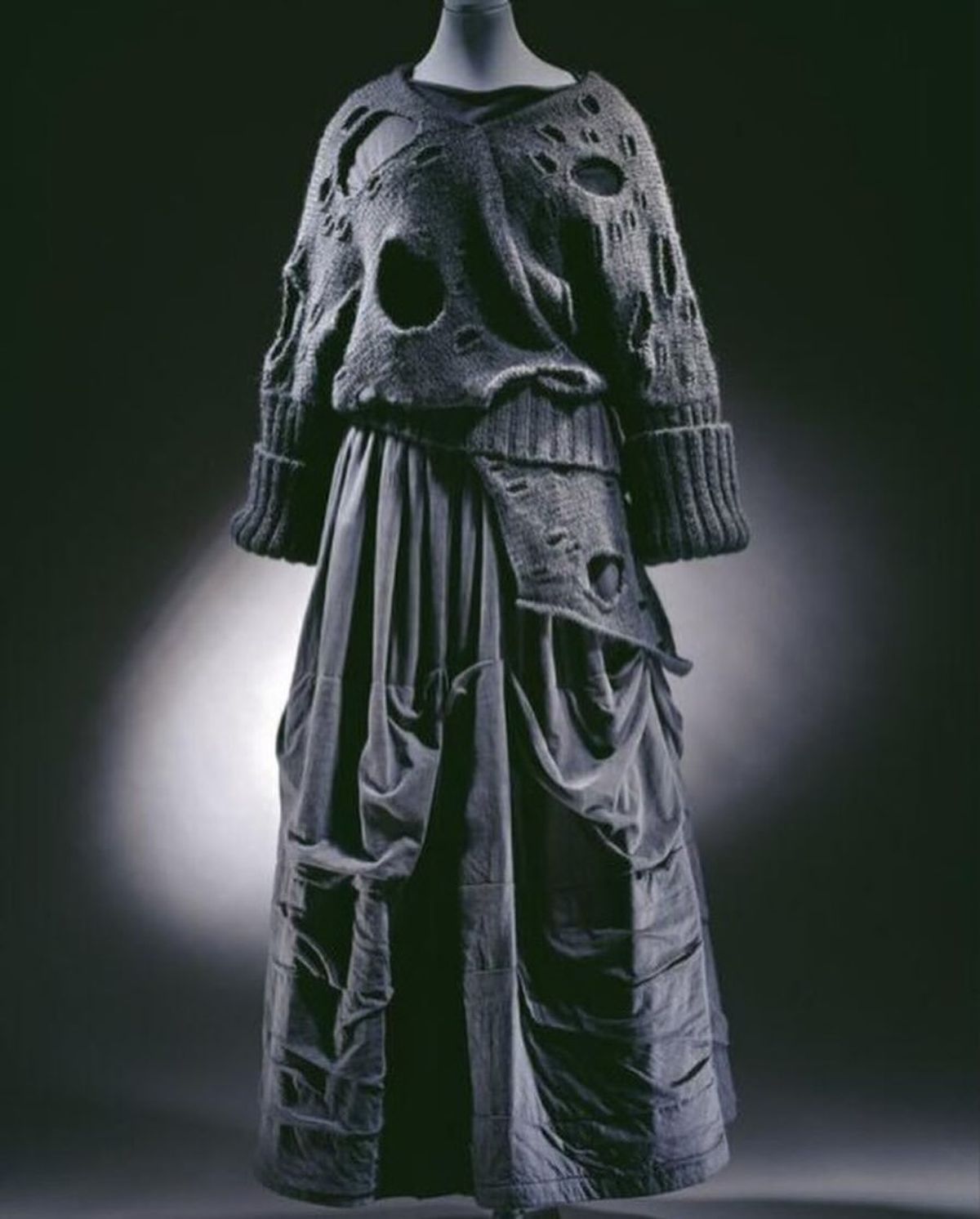 A mannequin lit against black in a grey knitted top full of large open holes, worn over a heavily gathered and padded long skirt.
