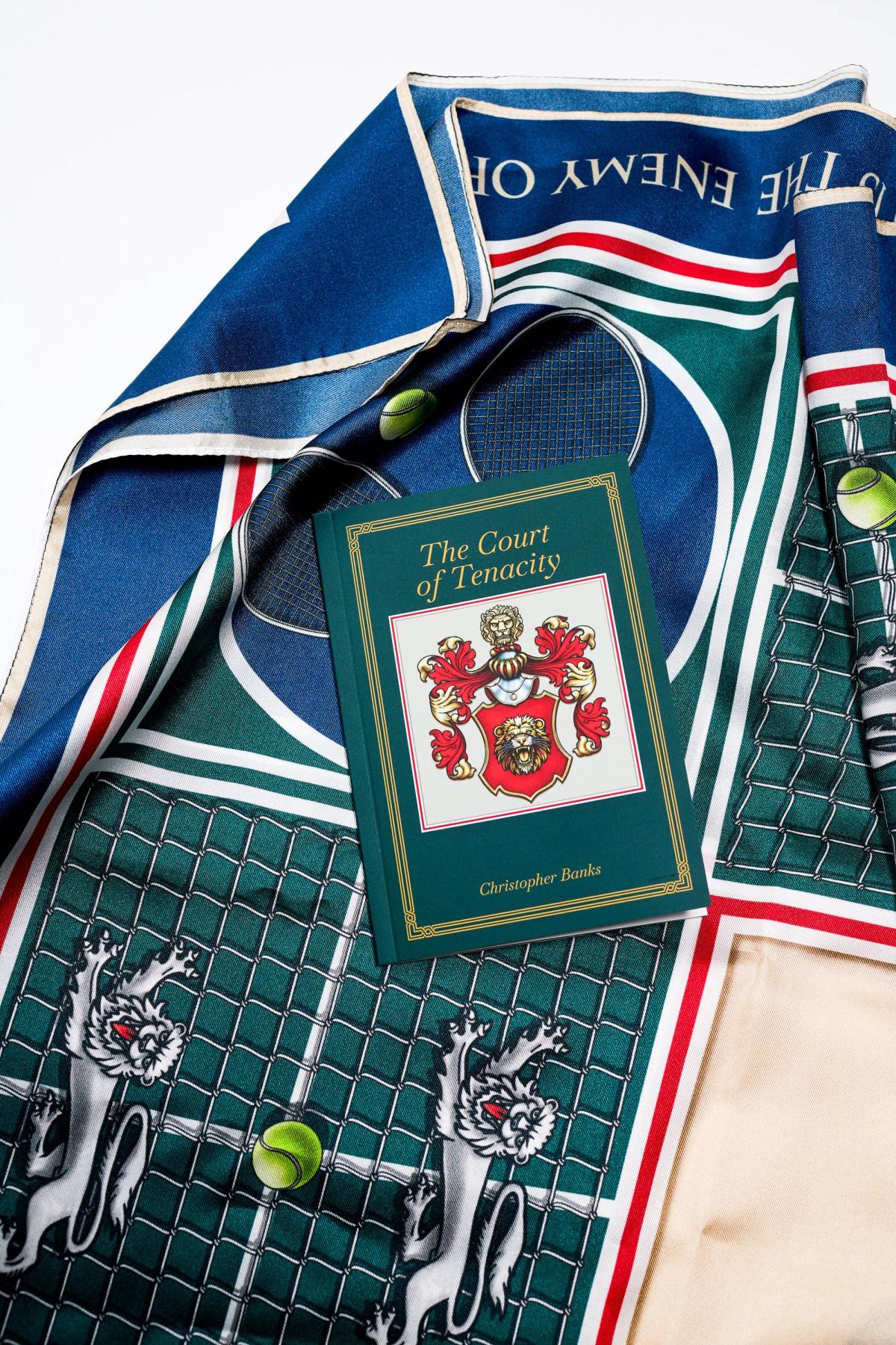 The Court of Tenacity book resting on the folded scarf, the crest on its cover echoing the printed silk beneath it.