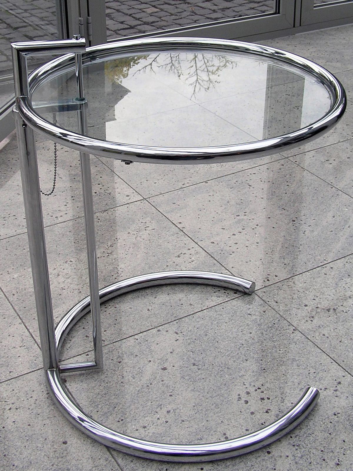 A circular glass table on a tubular chrome frame, its height adjustable, standing on a plain floor.