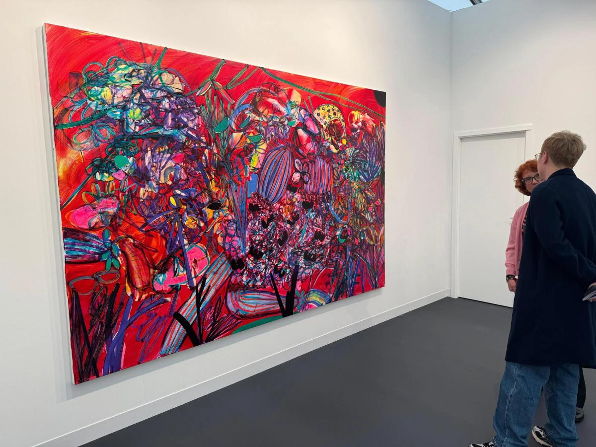 A large brightly coloured abstract painting on a white gallery wall, two visitors standing to one side looking at it.
