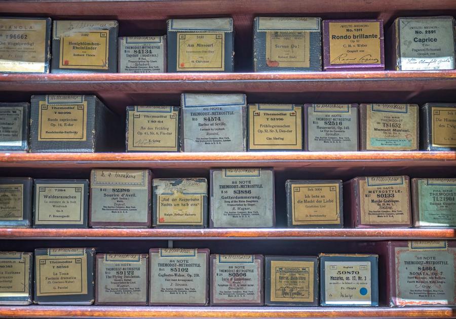 Wooden shelves holding rows of labelled piano roll boxes, each printed with a title, a catalogue number and a composer.
