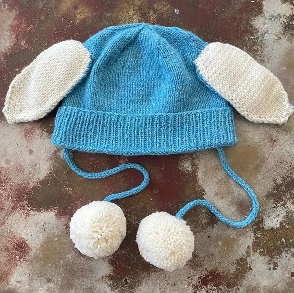 A pale blue hand knitted hat with long white knitted ears and two cream pompoms on cords, laid flat on a rusted metal surface.