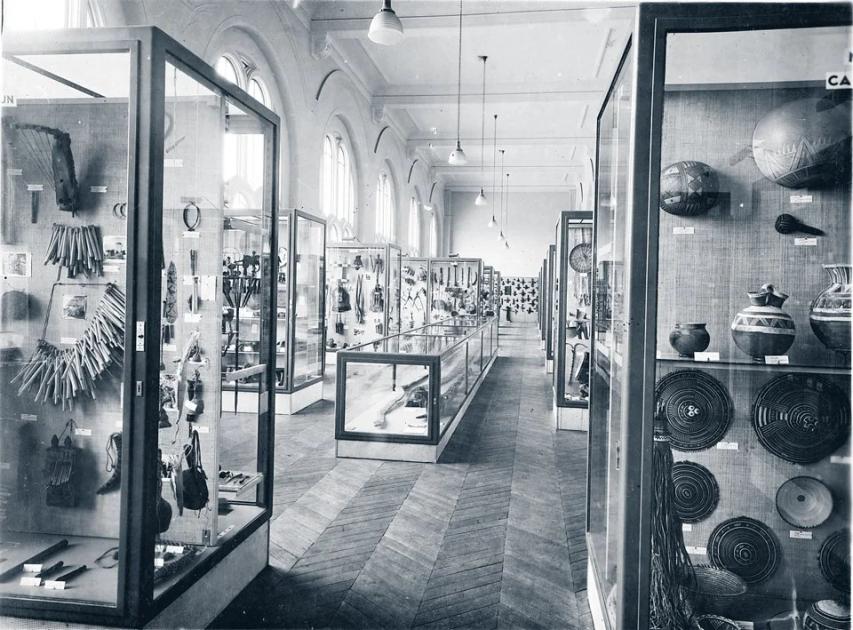 A black and white photograph of a museum gallery lined with tall glass cases holding baskets, pots and carved objects, receding down a long hall.