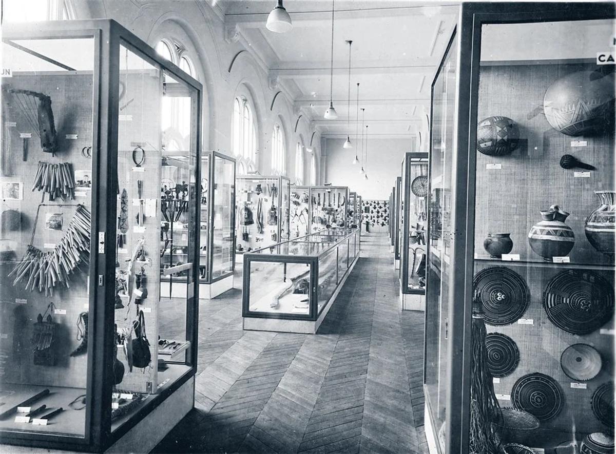 A black and white photograph of a museum hall lined with tall glass cases holding baskets, pots and carved objects receding down the room.