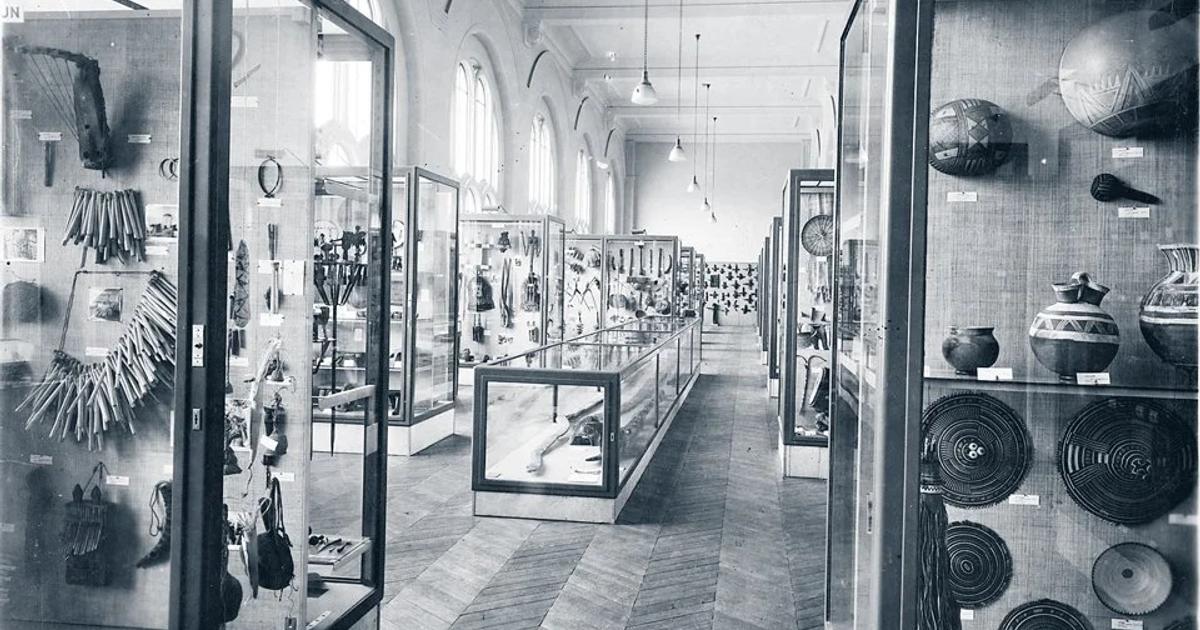 A black and white photograph of a museum gallery lined with tall glass cases holding baskets, pots and carved objects, receding down a long hall.