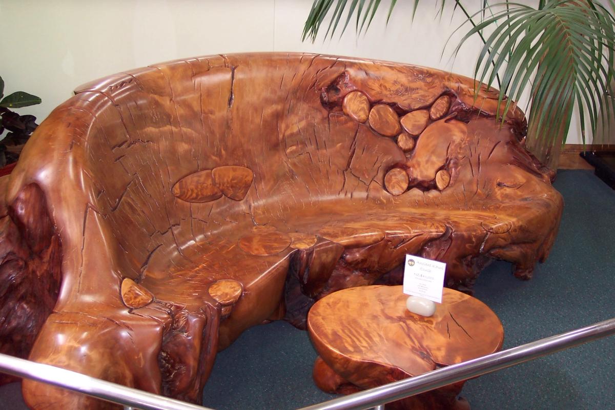 A large polished bench and small round table carved from a single piece of swamp kauri, displayed behind a rail in a museum, with a small printed card beside them.