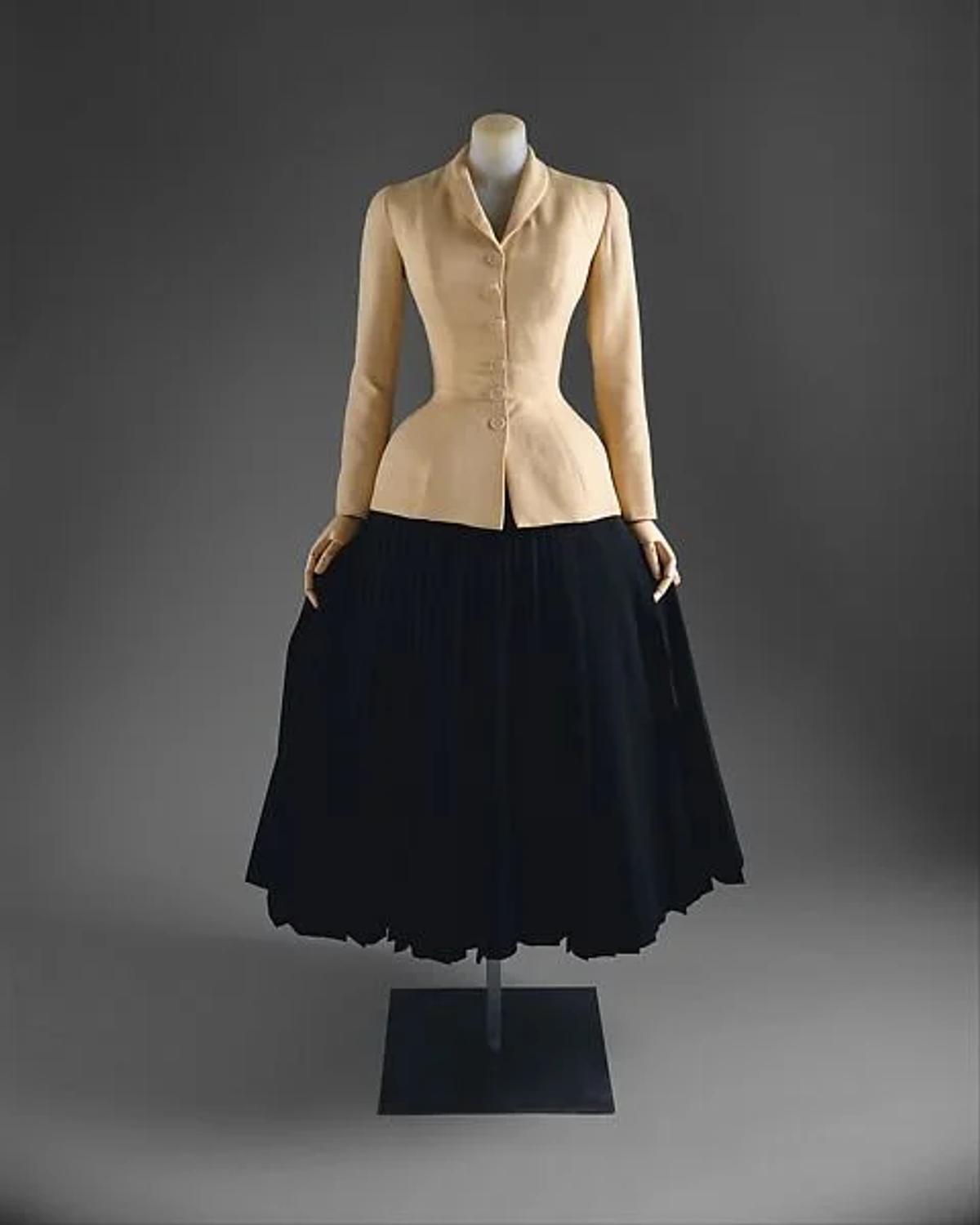 A tailored jacket with a tightly nipped waist in pale fabric shown on a mannequin above a full pleated black skirt.