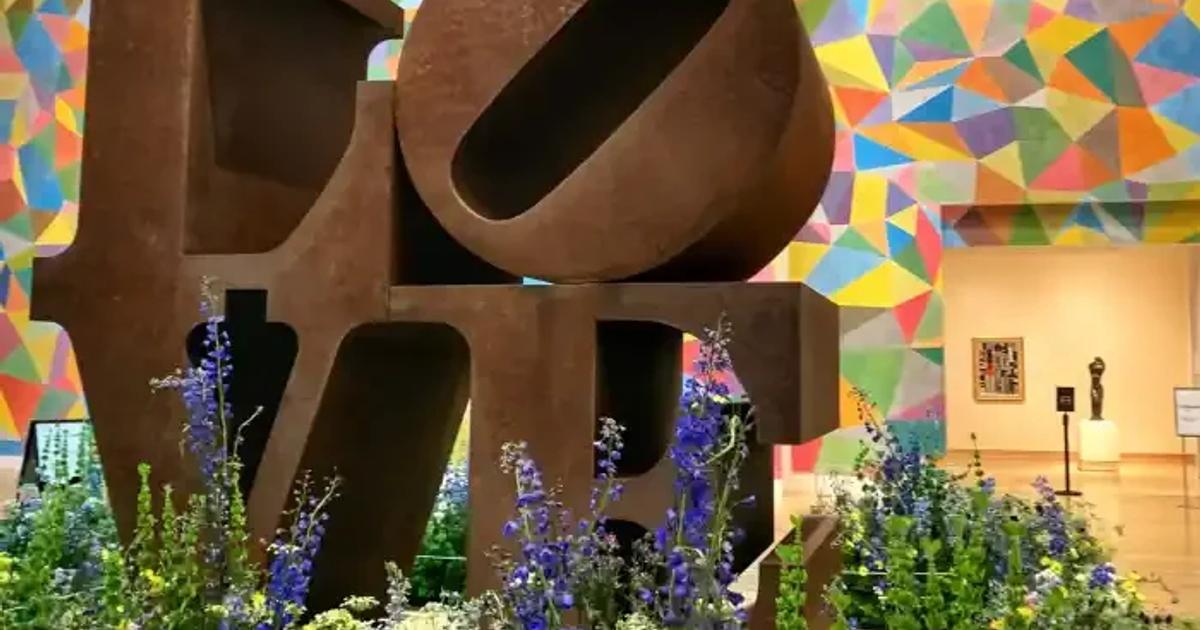 A weathered steel sculpture spelling the word love in stacked letters, set in a bed of flowering plants in front of a brightly coloured geometric mural.
