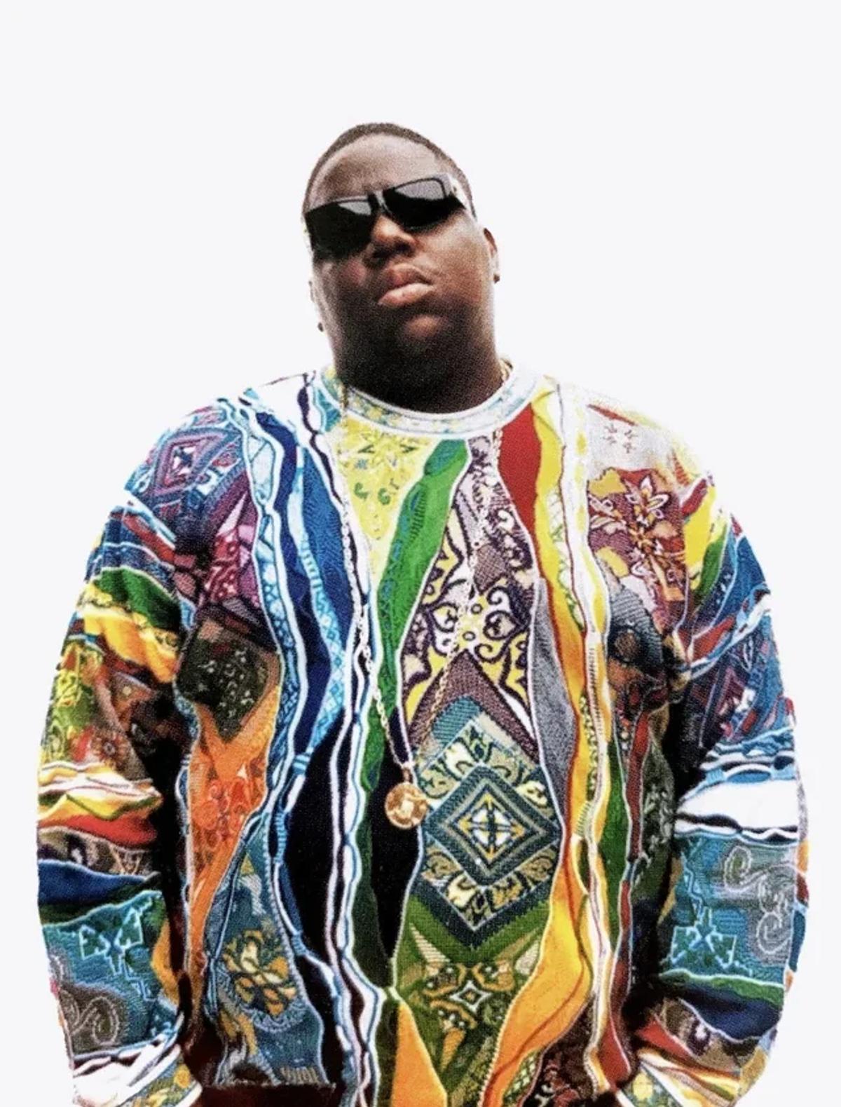 A man in dark sunglasses and a gold chain standing against a white background in a heavily patterned knitted sweater in many colours.
