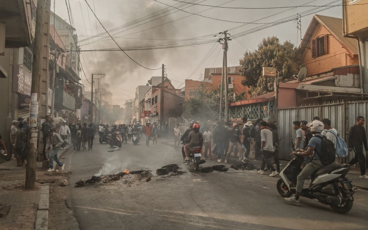 A street in Antananarivo with smoke rising from burning material laid across the road, people walking and riding motorcycles past it between low buildings and power lines.