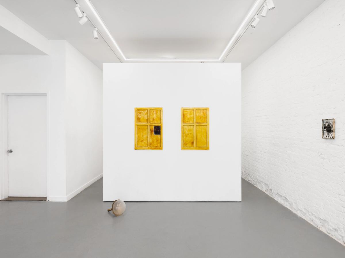 Two window-shaped glass panels in amber and yellow, each divided into four panes, mounted on a freestanding white wall. One pane holds a small dark photograph. A glass sphere rests on the floor below.