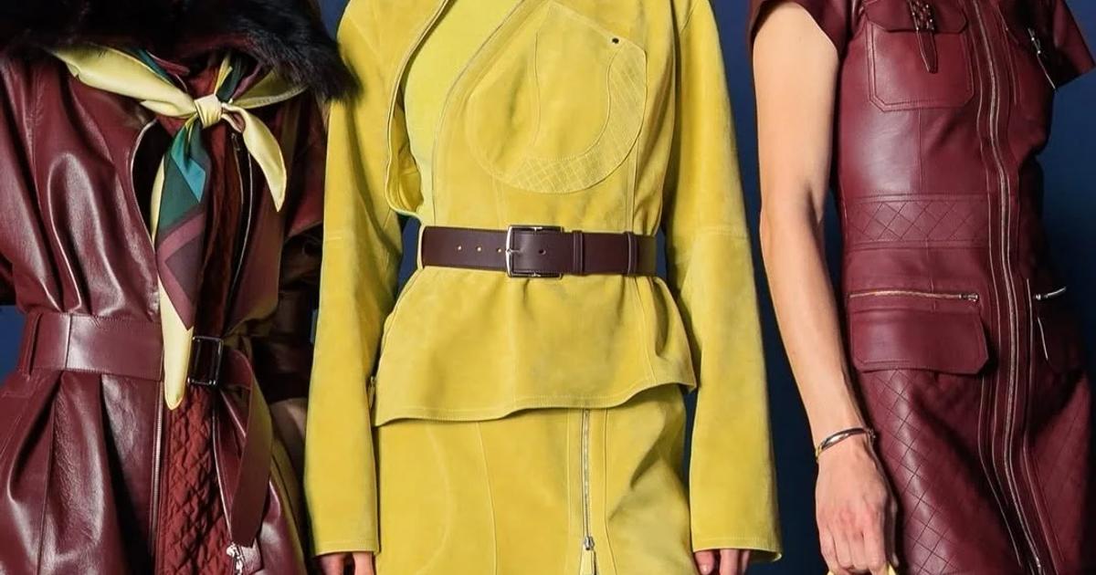 Three models standing against a dark blue backdrop in leather: an oxblood trench with a silk scarf, a belted chartreuse suede jacket and skirt, and a burgundy zipped dress with a small yellow handbag.