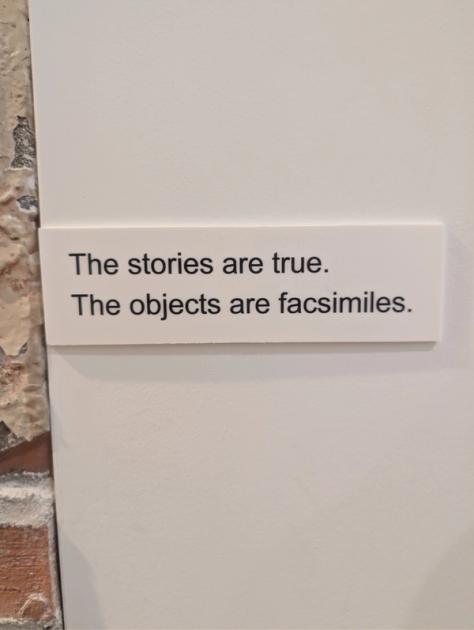 A small white label mounted on a gallery wall, printed in plain dark type with two sentences: The stories are true. The objects are facsimiles.