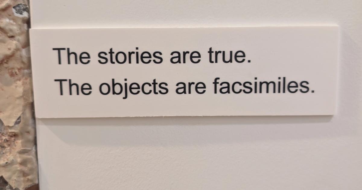 A small white label mounted on a gallery wall, printed in plain dark type with two sentences: The stories are true. The objects are facsimiles.