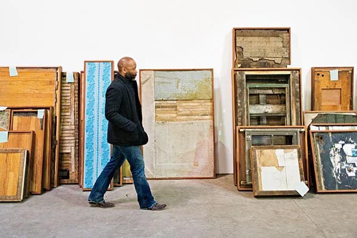 A man walking past a wall stacked with salvaged painted boards and framed panels leaning in rows inside a large workshop.