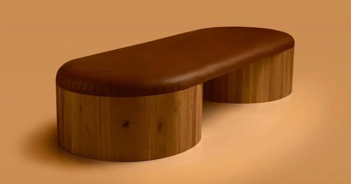 A solid timber bench formed from two rounded blocks joined by a lower curved seat, photographed against a flat orange background.