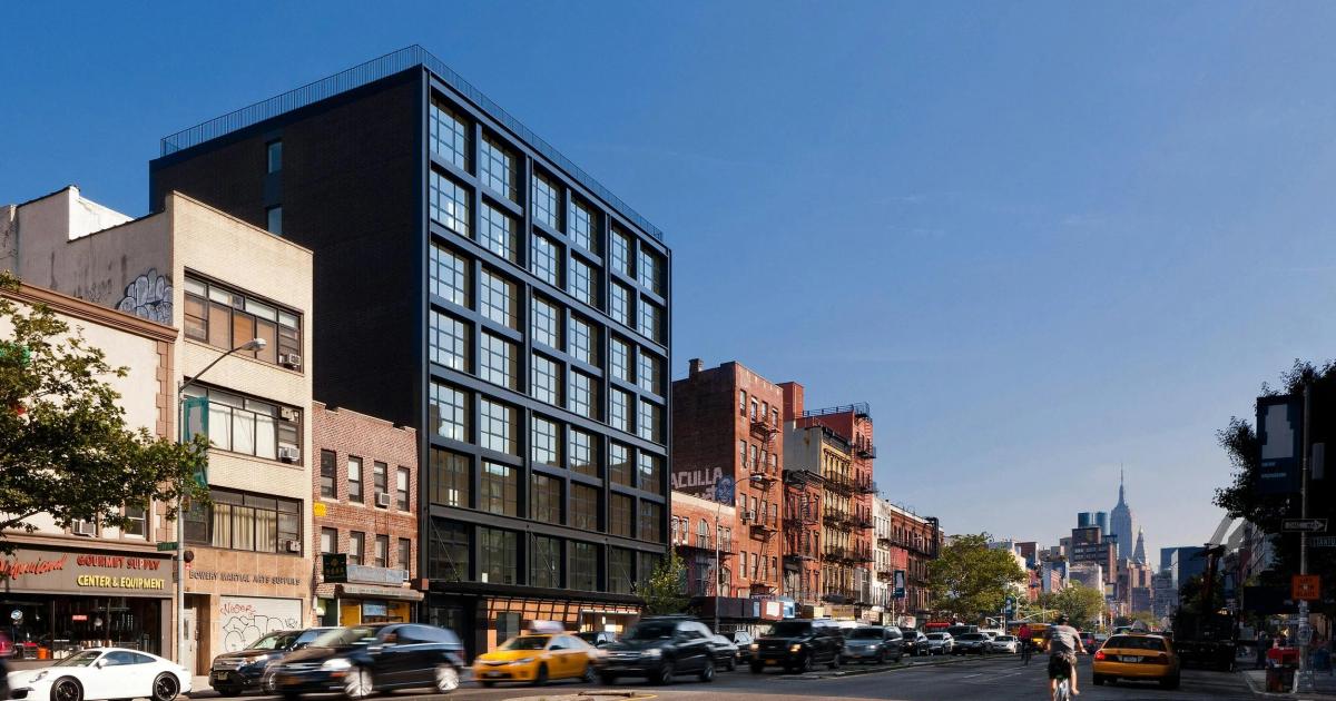 A dark modern gallery building mid block on a sunlit New York avenue, flanked by older brick and cast iron neighbours, traffic passing and the Empire State Building distant.