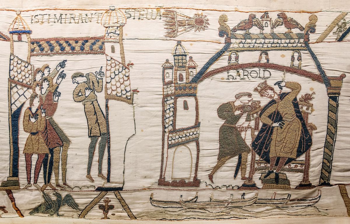 An embroidered linen panel showing robed figures pointing upward at a comet, a tower, and a seated crowned figure, with Latin lettering above.