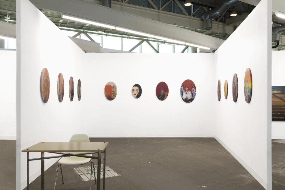 An empty art fair booth with white walls hung with a row of small round paintings, a folding table and chair standing on the concrete floor.