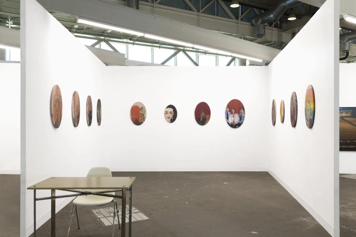 An art fair booth with white walls hung with a row of small round paintings, a folding table and chair on the concrete floor.