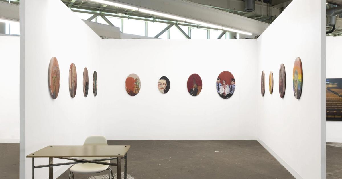 An empty art fair booth with white walls hung with a row of small round paintings, a folding table and chair standing on the concrete floor.