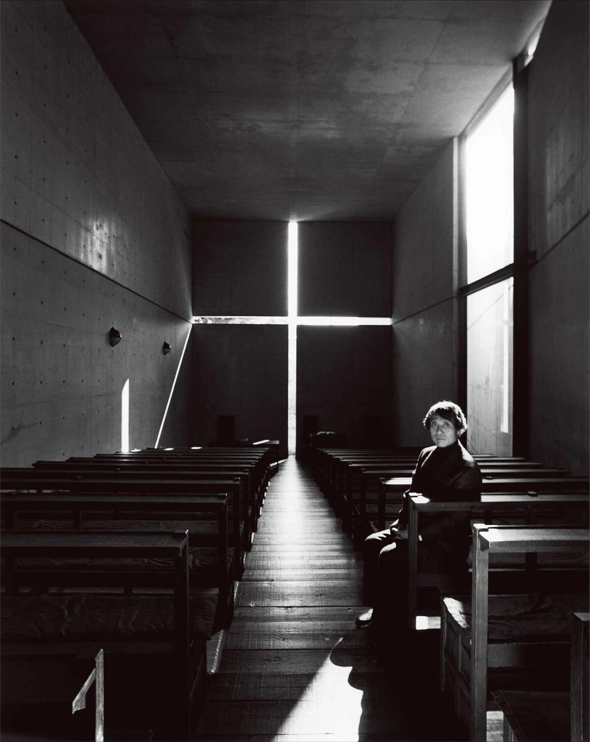 The interior of a concrete chapel where a cross shaped opening in the end wall admits bright daylight, with dark timber pews and a single seated figure.