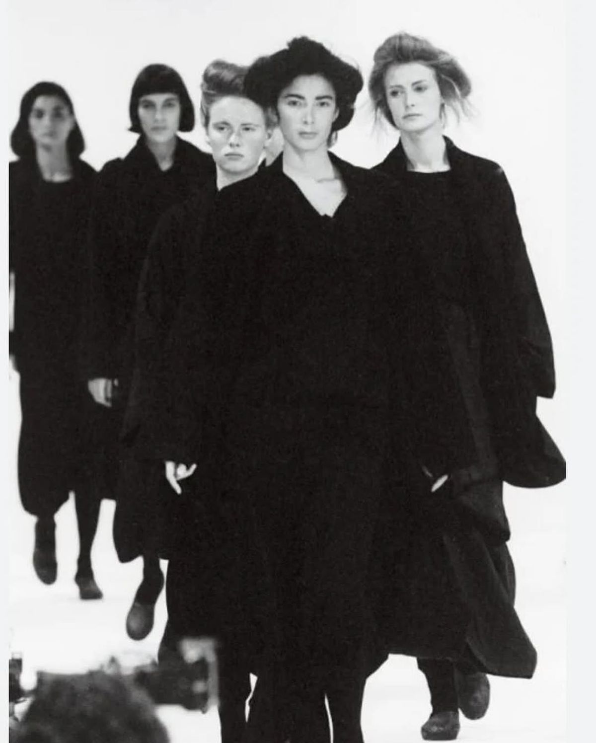 A black and white runway photograph of models walking forward in oversized black layered coats and knitwear, their hair cropped or pulled back.