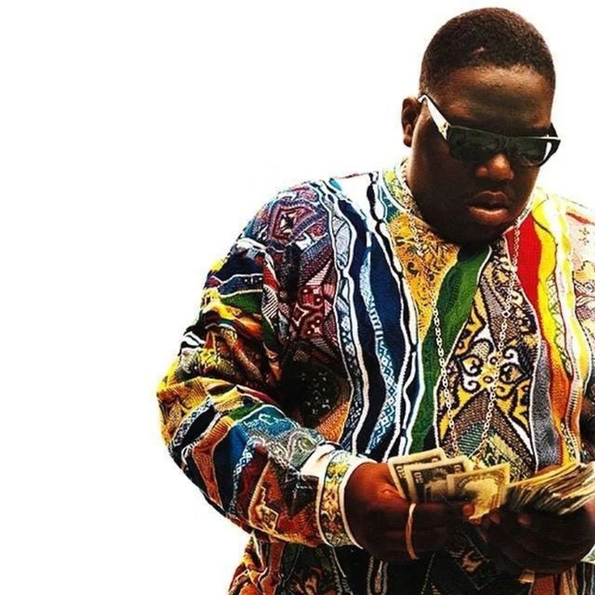 A man in dark sunglasses and a brightly patterned knitted sweater standing against a white background, counting a handful of banknotes.