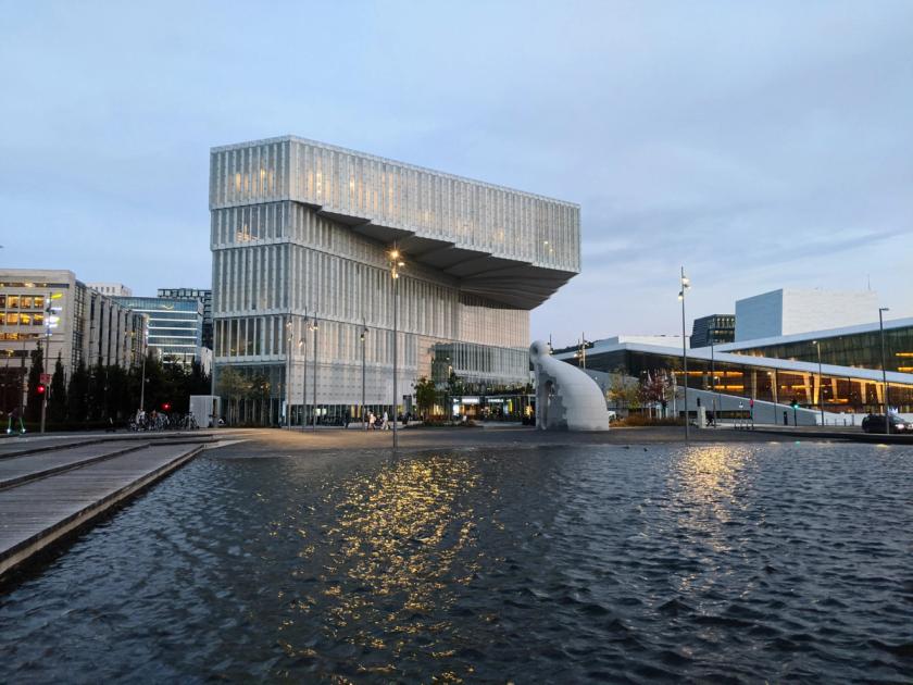 A low modern library building in pale cladding beside a shallow water mirror, with part of a white sloping opera house at right.