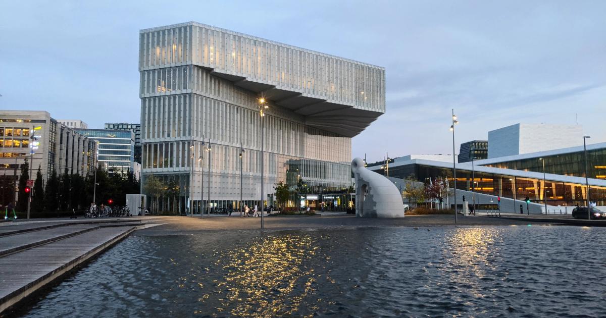 A low modern library building in pale cladding beside a shallow water mirror, with part of a white sloping opera house at right.