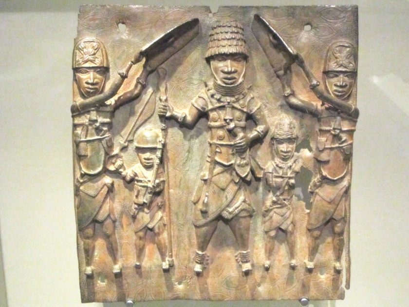 A cast brass relief plaque showing two warriors flanking and shielding a central figure, displayed in a museum case.