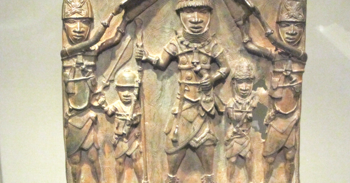 A cast brass relief plaque showing two warriors flanking and shielding a central figure, displayed in a museum case.