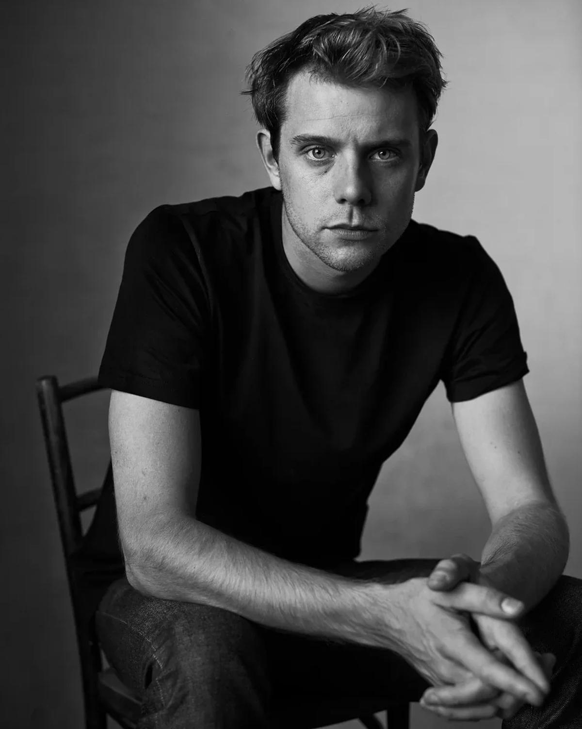 A black and white portrait of a young man in a plain dark t shirt seated on a chair with his hands clasped in front of him.