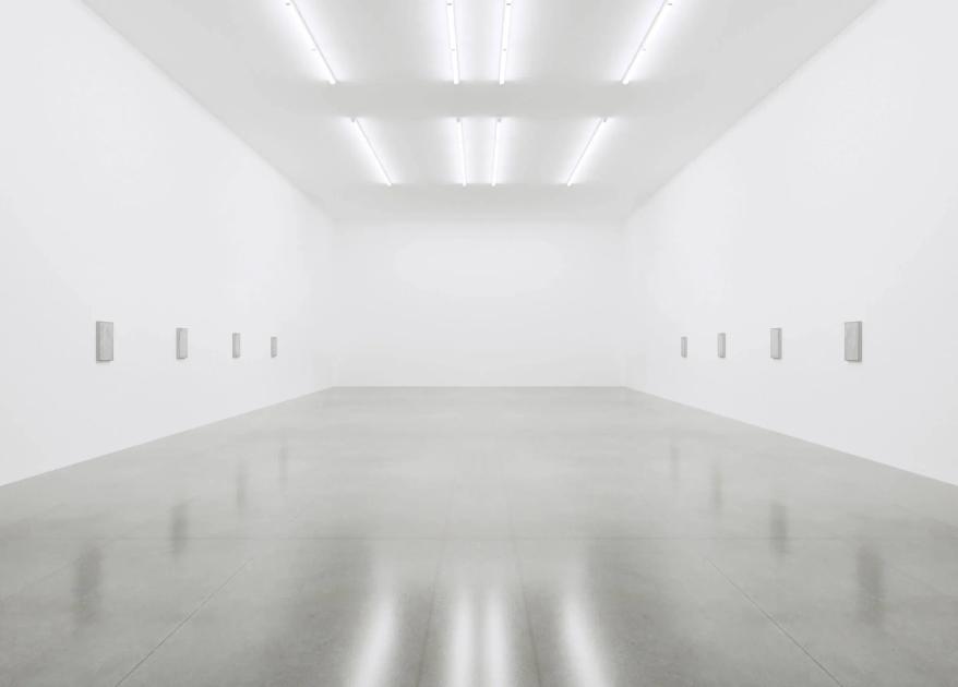 An empty white gallery room lit by ceiling strip lights, its polished floor reflecting the walls, with small blank labels spaced along both sides.