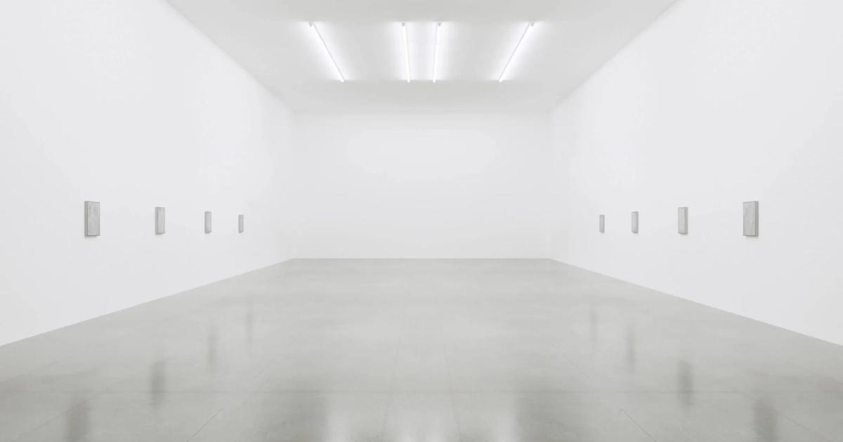 An empty white gallery room lit by ceiling strip lights, its polished floor reflecting the walls, with small blank labels spaced along both sides.