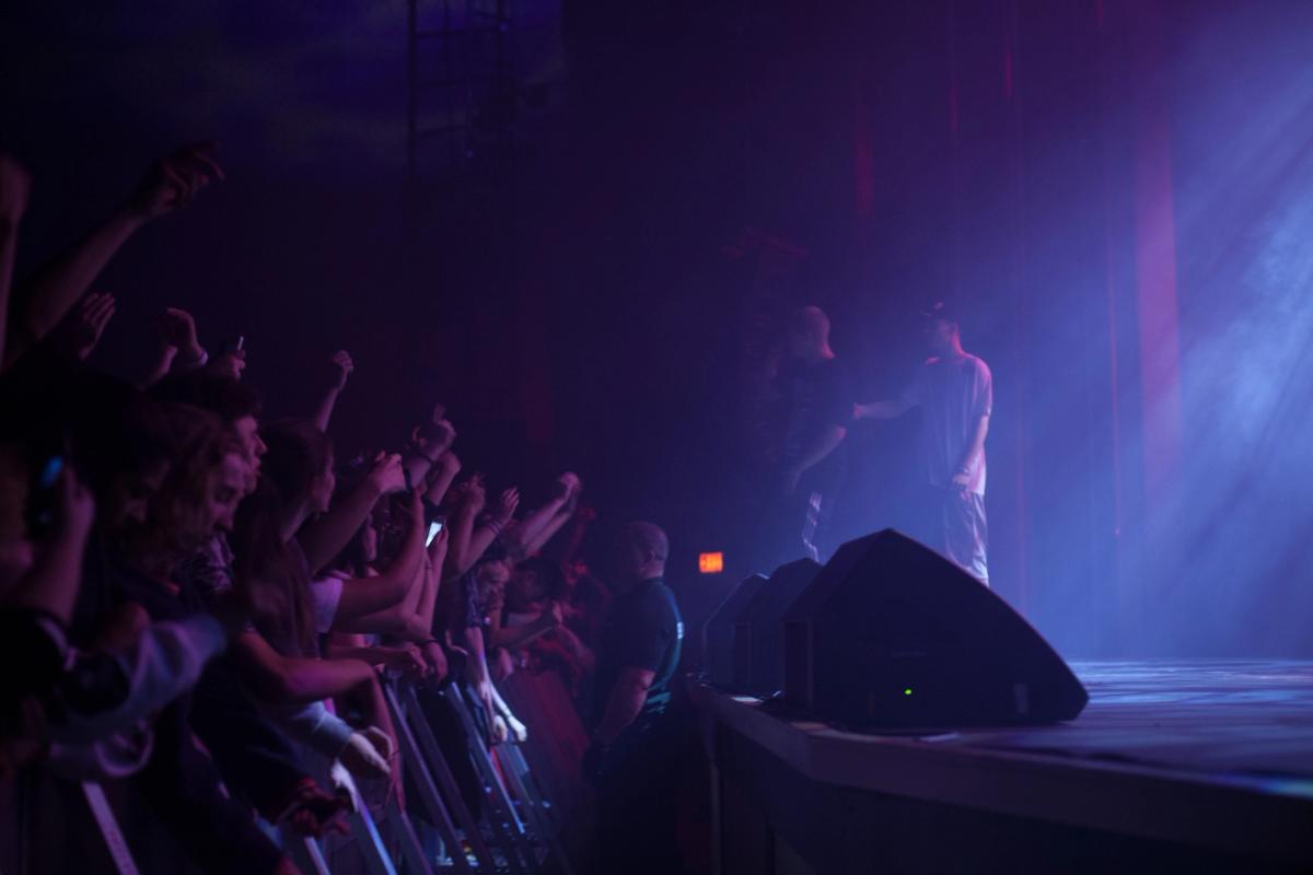 A crowd with raised arms at the barrier of a smoke-filled venue, the performer small and distant on stage.