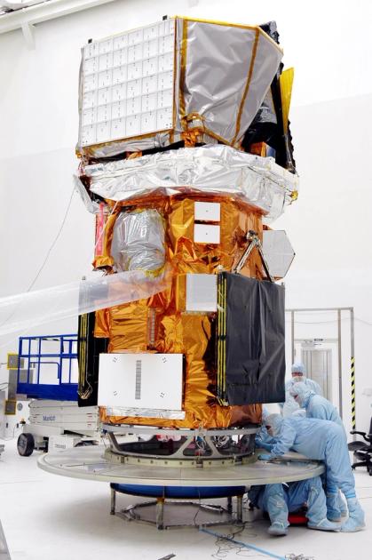 The Swift spacecraft upright in a cleanroom before launch, wrapped in gold and silver thermal blanketing, with technicians in blue coveralls crouched at its base.