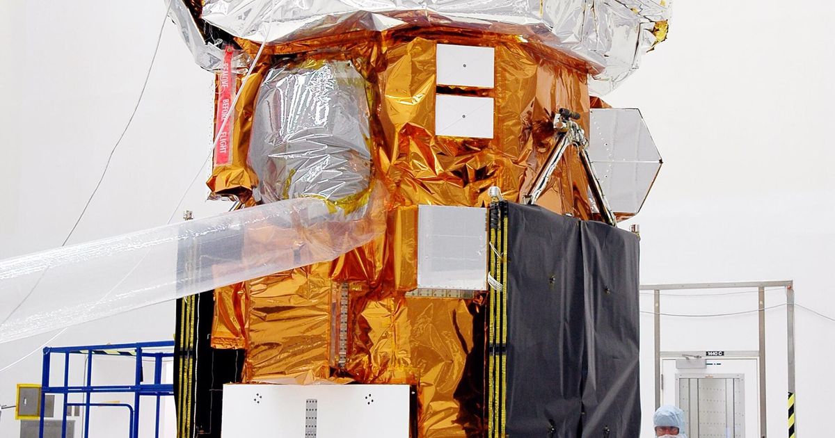 The Swift spacecraft upright in a cleanroom before launch, wrapped in gold and silver thermal blanketing, with technicians in blue coveralls crouched at its base.