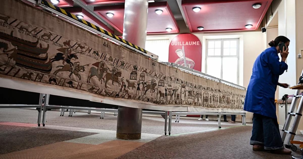 The Bayeux Tapestry laid out along low tables in a red ceilinged room, its embroidered ships and horsemen visible, with a woman in a blue coat standing at the far end.