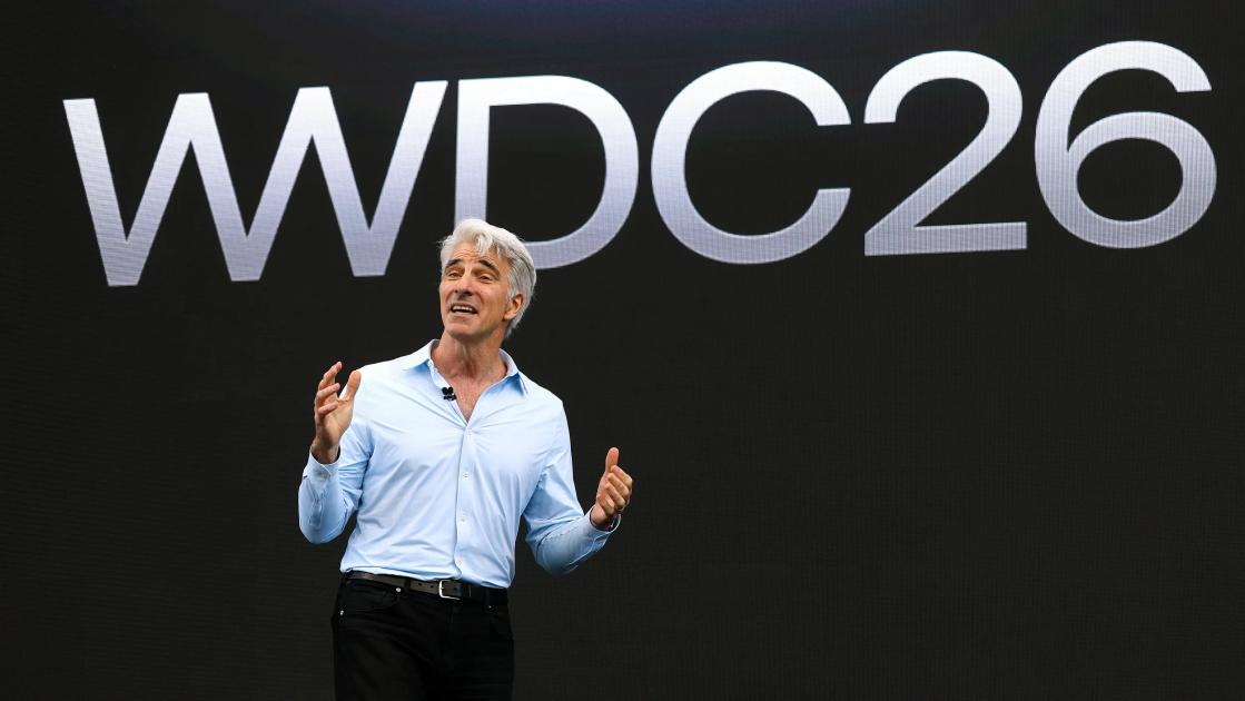 A man in a pale blue shirt speaking on a dark stage in front of a large screen reading WWDC26.