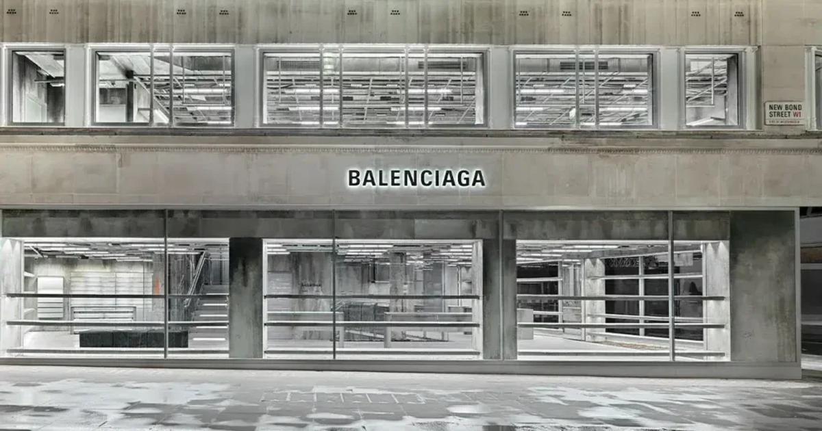 A black and white photograph of a modernist shopfront with continuous glazing and Balenciaga lettering above the windows, seen across an empty pavement.