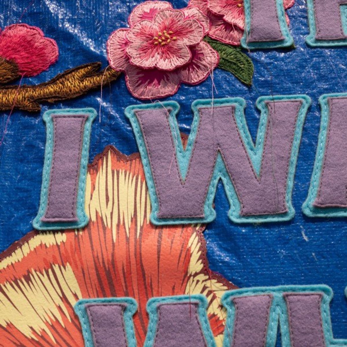 A detail of the embroidered banner showing raised pink blossom worked in thread over a blue ground beside pale appliqued capital letters.