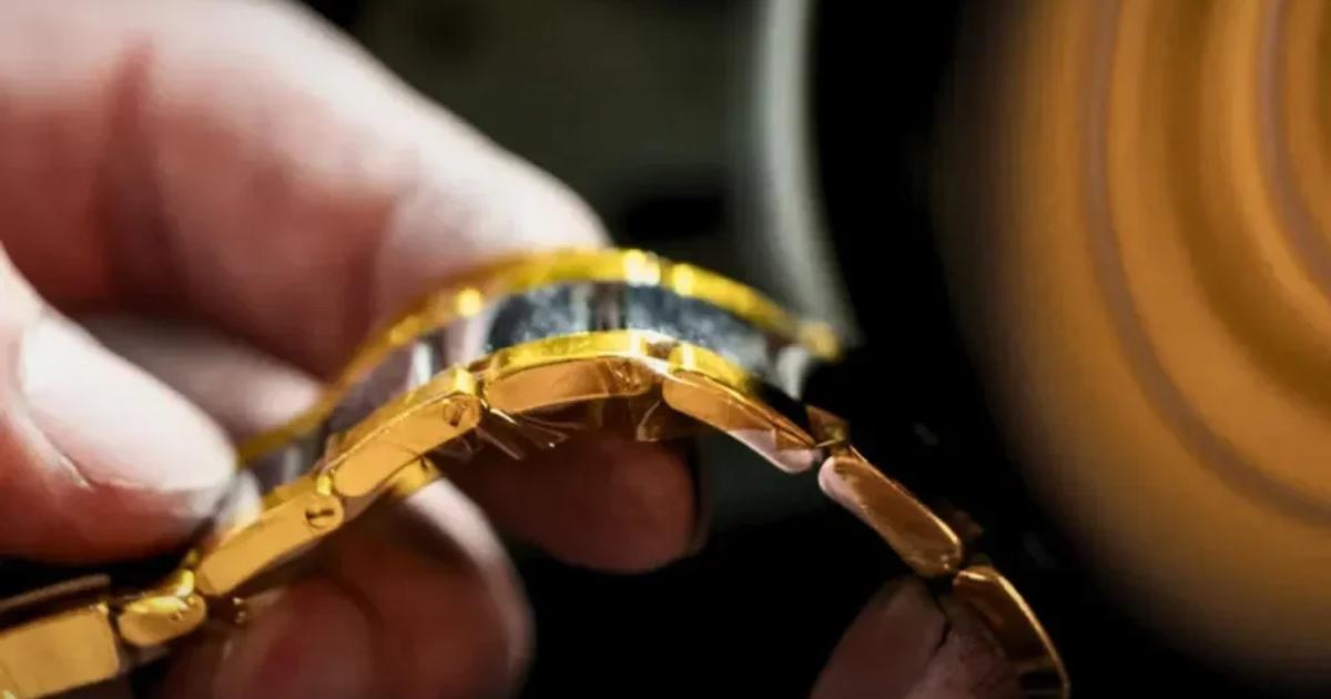 A close view of two hands holding a partly assembled gold watch bracelet at a workbench, a polishing wheel spinning out of focus behind them.