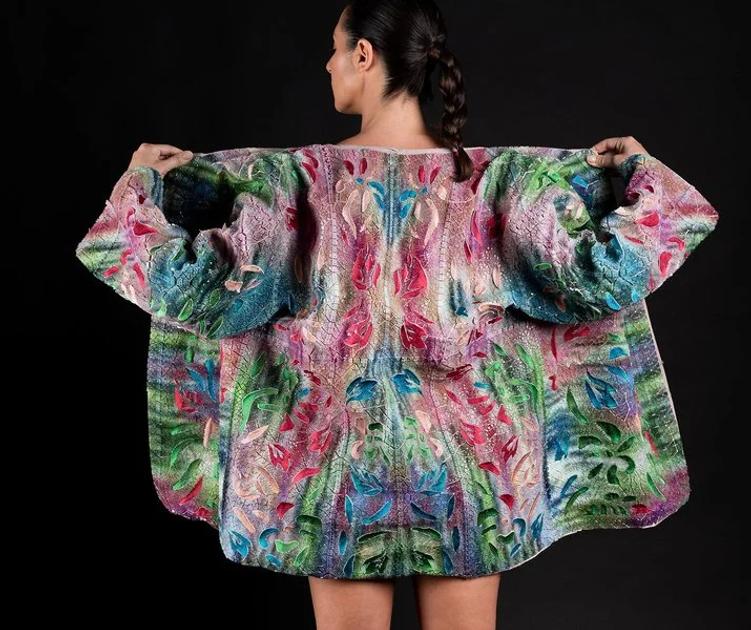 The back of a woman wearing an oversized knitted coat in pink, green and blue, its pattern mirrored down the spine, arms held wide against a black background.