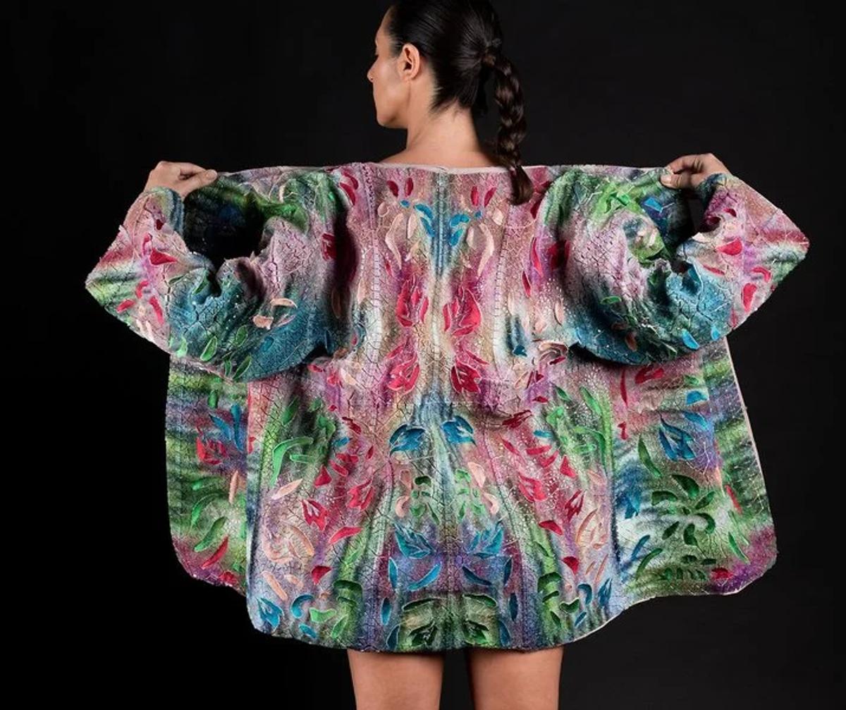 The back of a woman wearing a wide oversized coat woven in swirling pink, green and blue, the pattern mirrored down the spine, against black.