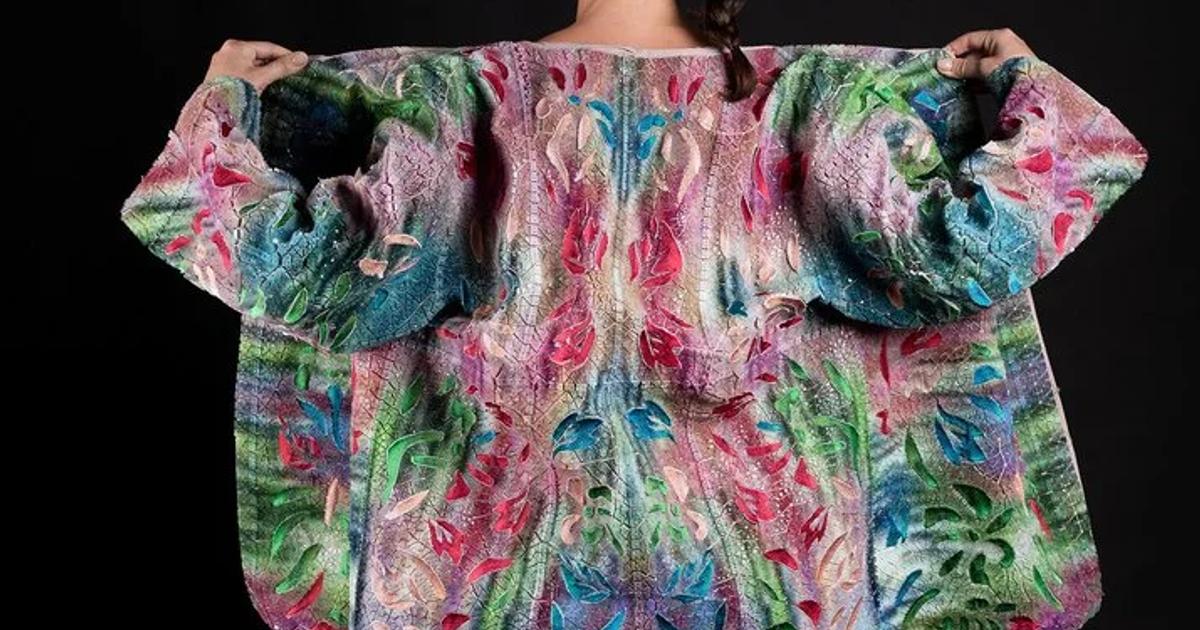 The back of a woman wearing an oversized knitted coat in pink, green and blue, its pattern mirrored down the spine, arms held wide against a black background.
